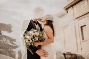 A couple on their wedding day cuddles together while kissing while the veil covers both of them and the bride holding her bouquet.