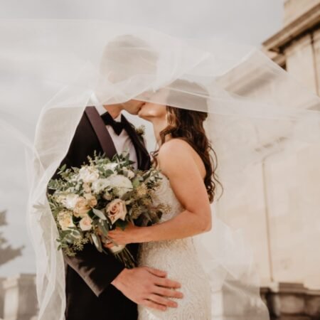 A couple on their wedding day cuddles together while kissing while the veil covers both of them and the bride holding her bouquet.