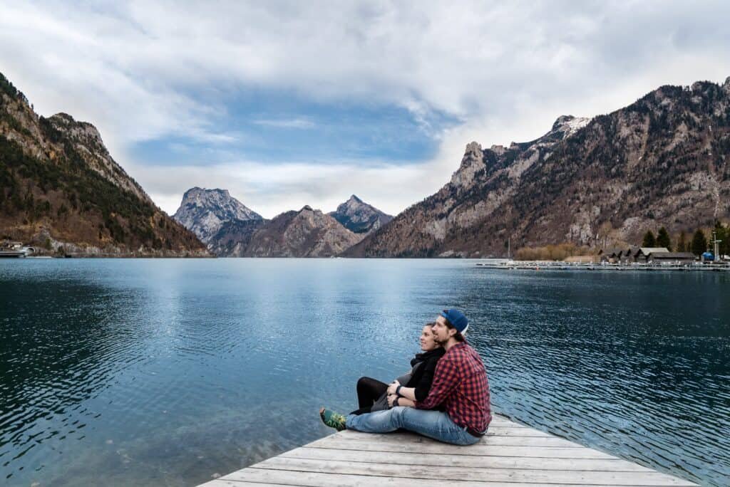 A young couple seated and cuddling in a board by a river while taking in views of gorgeous mountains nearby.