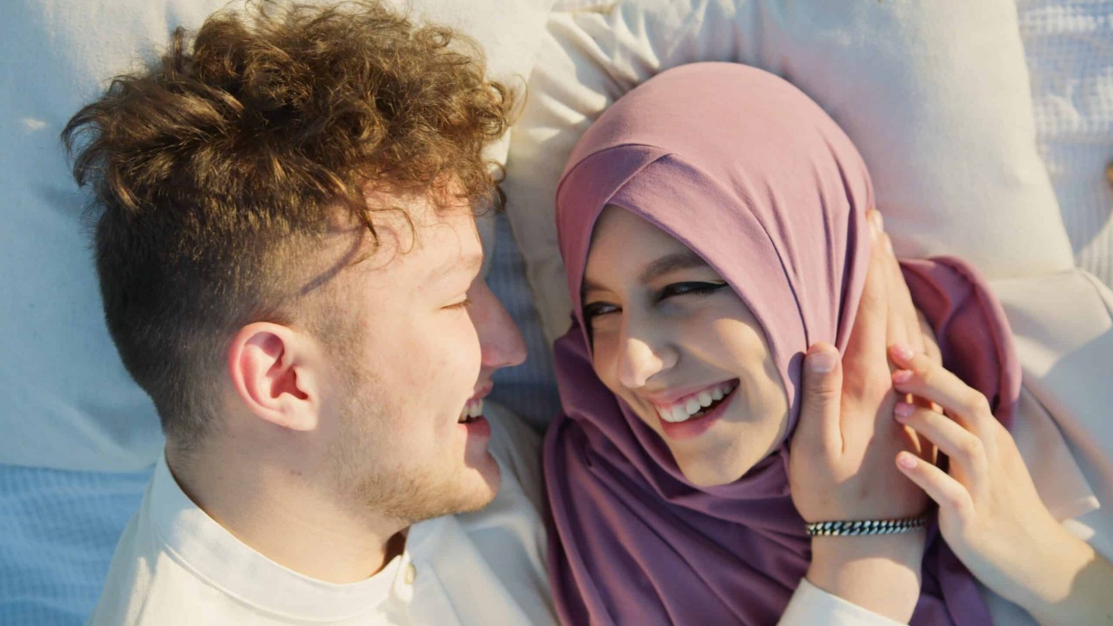 A young Muslim couple is smiling, holding hands and staring into their eyes while in bed, happy about speaking their love language of quality time.