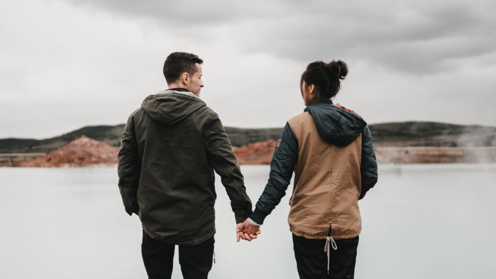 A couple is holding hands while walking towards a lake and hillside.