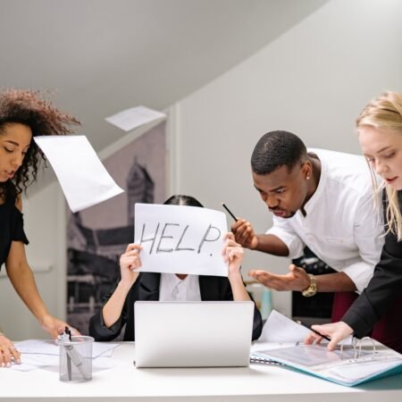 A lady displaying the "help" sign while being swamped by three workmates with papers flying around her desk. Emotional intelligence 2.0