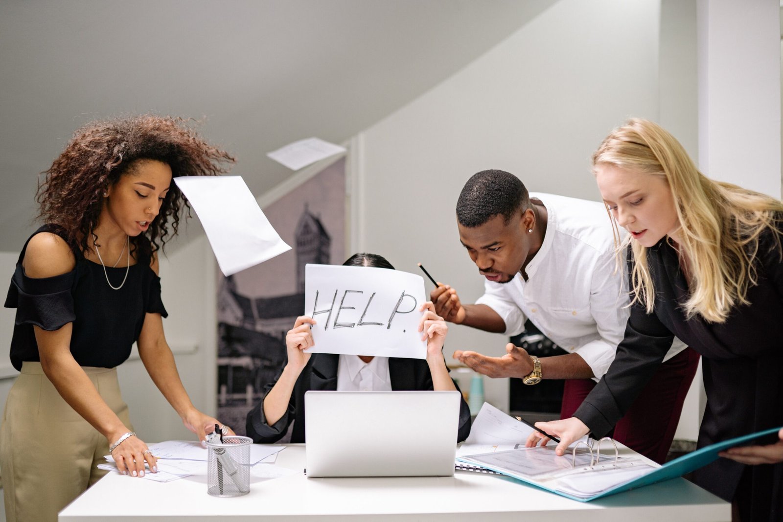 A lady displaying the "help" sign while being swamped by three workmates with papers flying around her desk. Emotional intelligence 2.0