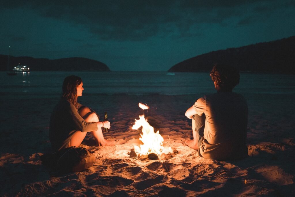 A couple hanging out by an open fire by the beach at dusk.