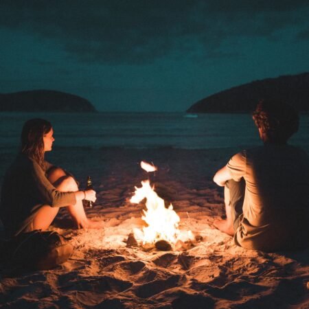 A couple hanging out by an open fire by the beach at dusk.