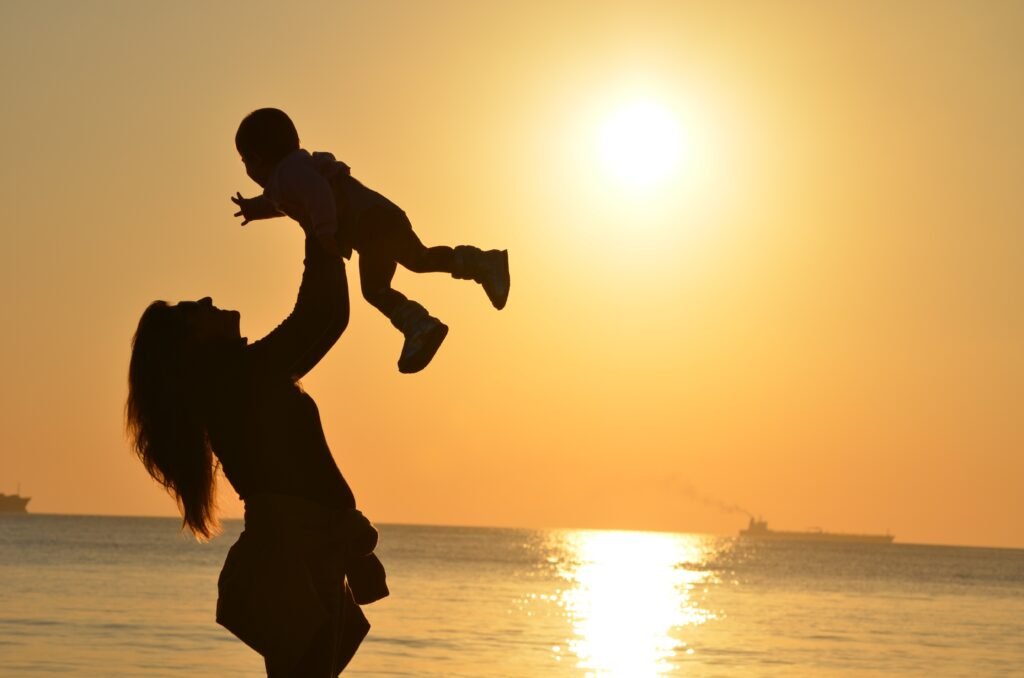 The silhouette of a mum lifting her baby up by the beach with the sunset at the backdrop.