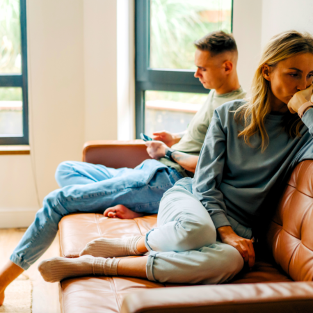 A sad wife looking away from her husband who is scrolling his phone as evidence of resentment in marriage.